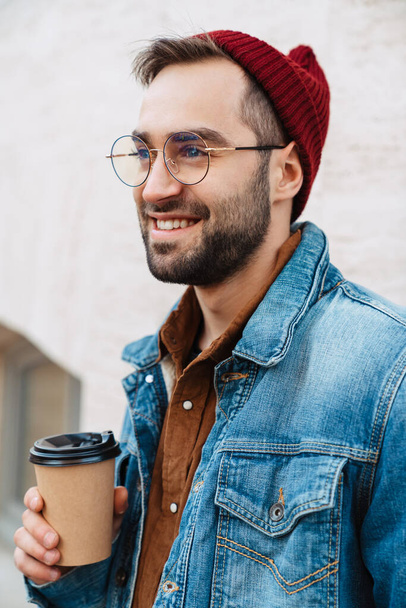 Close up of a handsome happy young stylish bearded man walking outdoors in the street, holding takeaway coffee cup - Foto, imagen