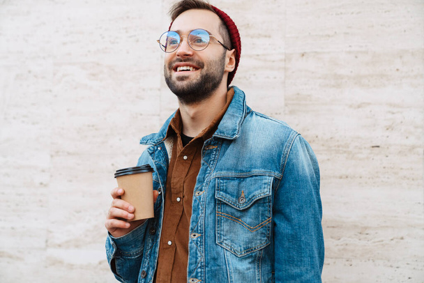 Close up of a handsome happy young stylish bearded man walking outdoors in the street, holding takeaway coffee cup - 写真・画像