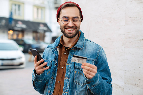 Close up of a handsome happy young stylish bearded man walking outdoors in the street, shopping online with mobile phone and credit card - Fotoğraf, Görsel
