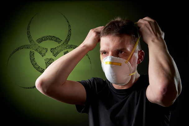 young man putting on a protective mask in dark atmosphere - black biohazard sign / emblem on green background - Zdjęcie, obraz