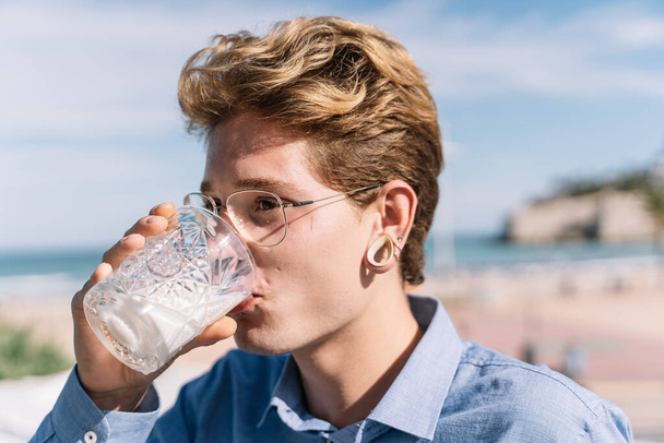 Niño rubio con gafas y piercings bebiendo agua en un vaso en la terraza de un restaurante - Foto, imagen