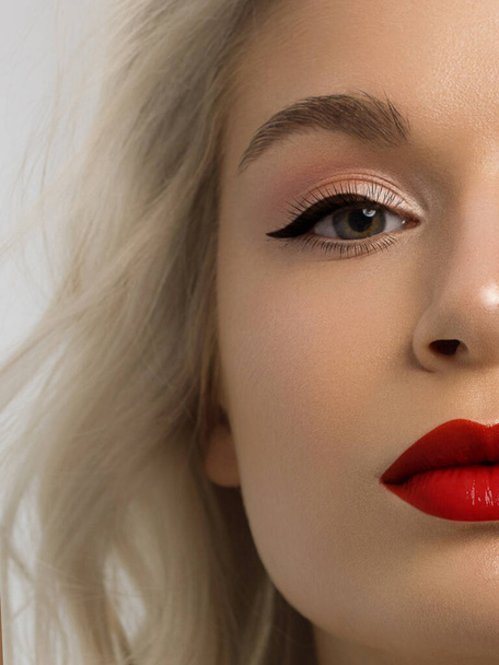 Close-up beauty of a half female face with creative fashion evening make-up. Black arrows on the eyes and extremely long eyelashes, on plump lips matte scarlet lip color. Well-groomed skin after spa - Фото, изображение