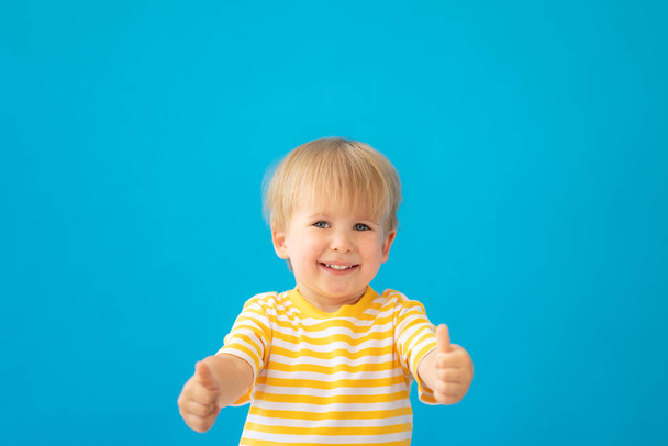 Close up portrait of happy child wearing striped shirt against blue background. Kid having fun on summer vacation. Boy showing thumbs up - Foto, afbeelding