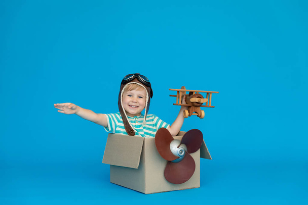 Happy child dreams of becoming a pilot. Kid having fun against blue paper background. Boy wearing striped shirt playing in cardboard box. Summer vacation and travel concept. Dream and imagination - Photo, Image