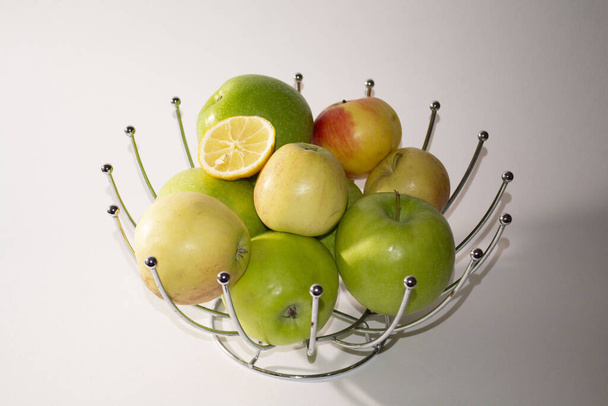 Yeşil sarı kırmızı elmalar ve metal dekoratif sepette bir dilim limon. Pencereden gelen ışık soldaki sepetin üzerine düşüyor. - Fotoğraf, Görsel