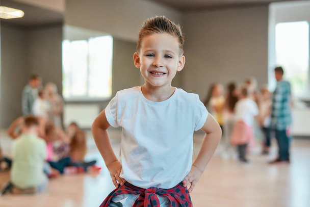 Cool dancer. Portrait of a cute and happy little boy in white t-shirt keeping arms on hips and looking at camera with smile while standing in the dance studio - Foto, Imagem