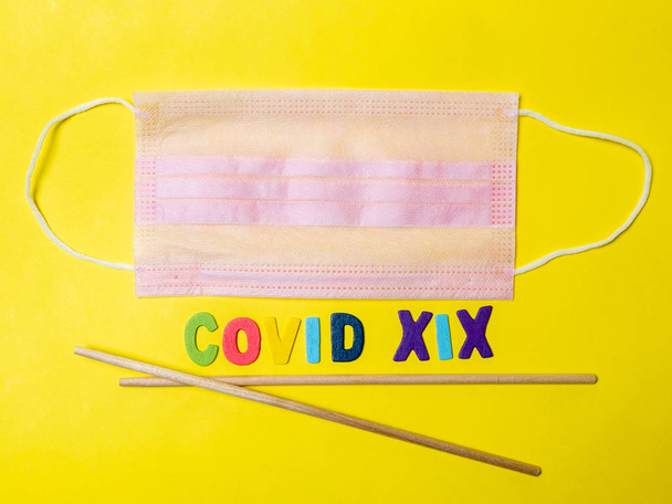 Pink medical mask on a yellow background and Asian chopsticks in the shape of the letter V. The inscription indicates the virus COVID-19. Disease as a treat from Asia. - Фото, зображення