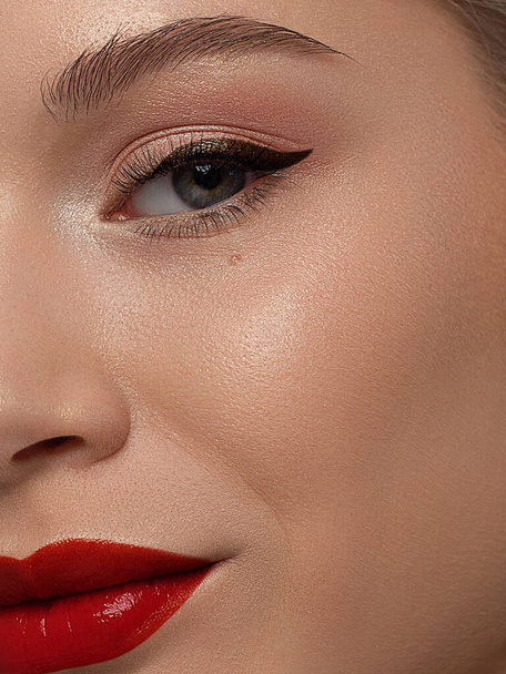 Close-up beauty of a half female face with creative fashion evening make-up. Black arrows on the eyes and extremely long eyelashes, on plump lips matte scarlet lip color. Well-groomed skin after spa - Photo, image