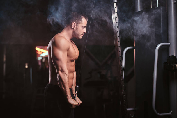 Ajuste deportista se concentró en un ejercicio en una máquina de tracción manual, bombeando sus tríceps en un gimnasio oscuro rodeado de humo
 - Foto, Imagen