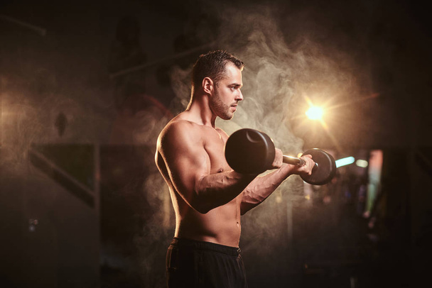 Shirtless bodybuilder doing weightlifts with barbell in a dark gym surrounded by smoke - Photo, image