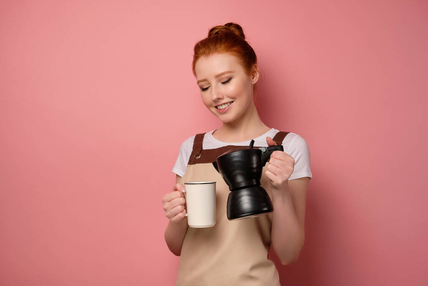 The red-haired girl in an apron stands in a half-turn on a pink background, tilting the coffee pot to the cup - Photo, image