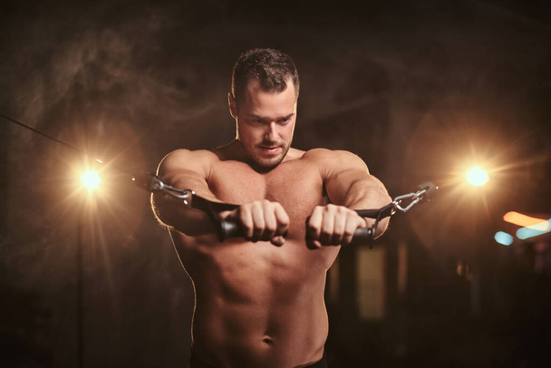 Спортсмен сближает руки, делая упражнения для грудных мышц в спортзале, окруженном дымом
 - Фото, изображение