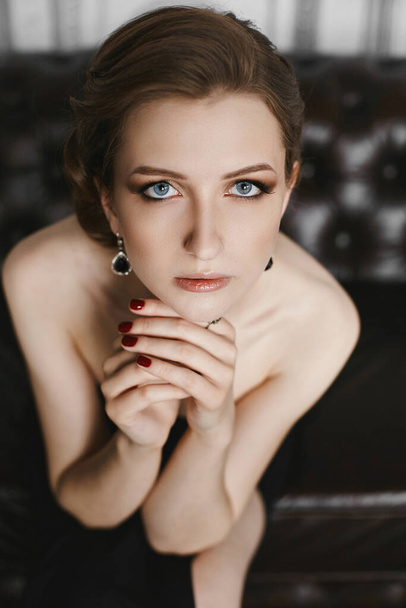 Portrait of elegant and beautiful young brunette model woman with gentle make-up, blue eyes and fashionable hairstyle, in long black dress sits on the vintage sofa at luxury interior - 写真・画像
