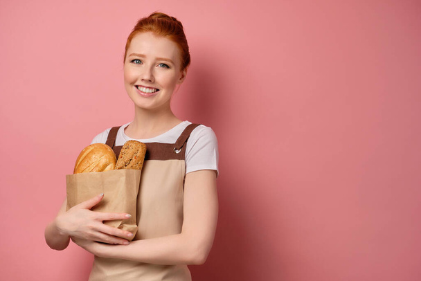 Red-haired girl in an apron, stands with a bag of bread on a pink background and smiles. - Фото, изображение