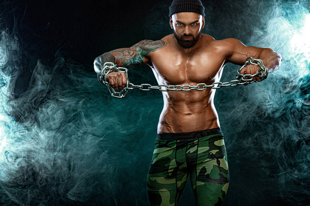 Athlete bodybuilder on black background with lights and smoke. Men fashion. Portrait of a brutal bearded man topless with chains. - Φωτογραφία, εικόνα