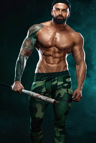 Strong and fit man bodybuilder with baseball bat shows abdominal muscles under a t-shirt. Sporty muscular guy athlete. Sport and fitness concept. Mens fashion. - Foto, Imagen