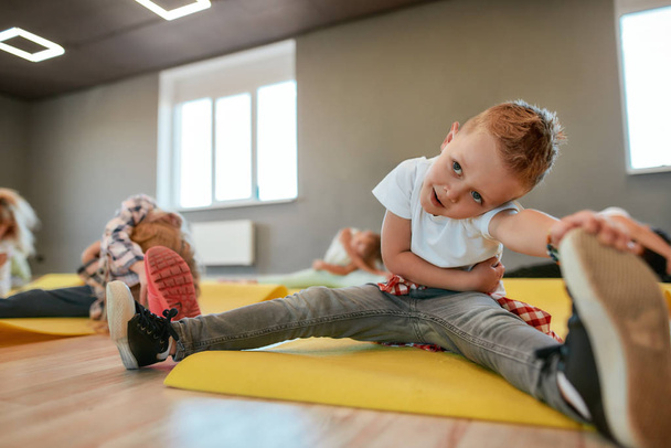 Close-up portret van een kleine jongen zittend op yoga mat en het strekken van zijn benen. Groep kinderen doet gymnastische oefeningen in de dansstudio - Foto, afbeelding