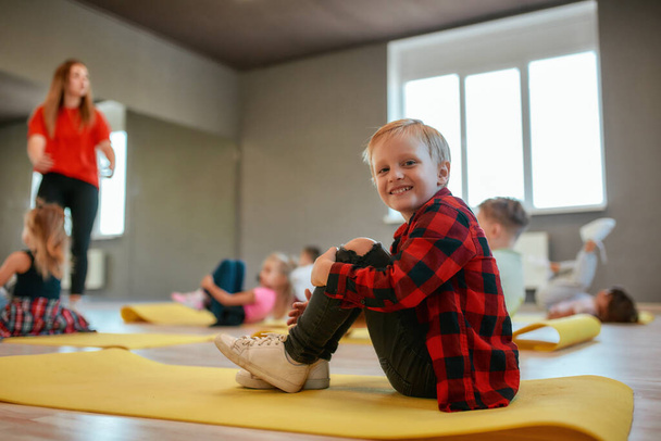 Positive little boy in casual clothes sitting on the mat and smiling at camera. Group of children doing exercises with female trainer in the dance studio - Foto, immagini