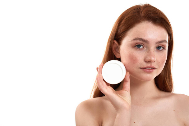Perfect face cream for her skin type. Young attractive red-haired girl with freckles demonstrating cosmetic product and looking at camera while standing in studio against white background - Foto, afbeelding