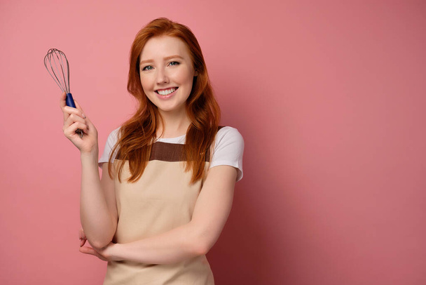 A beautiful red-haired girl in an apron stands on a pink background and smiles broadly at the camera holding a whisk in her hand - Photo, image