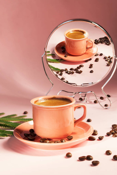 Korálově zbarvený šálek s kávovými zrnky se stíny, které se odrážely v zrcadle na pozadí pastelové růže. Koncept kávy. Letní zátiší, vertikální obraz - Fotografie, Obrázek