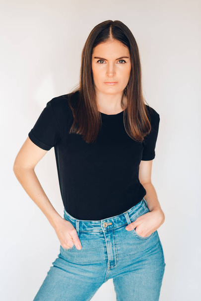 Indoor portrait of beautiful young woman, wearing black t-shirt and high waist jeans, posing on white background, holding hands in pockets - Foto, Imagem