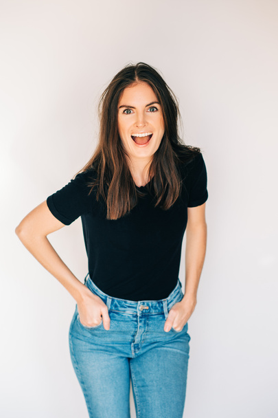 Indoor portrait of beautiful laughing woman, wearing black t-shirt and high waist jeans, posing on white background - Фото, изображение