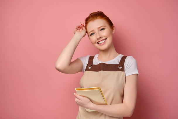 A red-haired girl in an apron with collected hair stands with a notepad and smiles broadly, putting away a pencil by the ear - Фото, изображение
