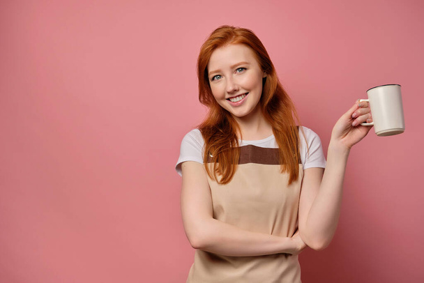 The red-haired girl in an apron stands on a pink background with a cup in her hand and smiles - Photo, image