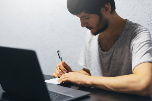 Portret młodego mężczyzny piszącego w notatniku, pracującego z laptopem. - Zdjęcie, obraz