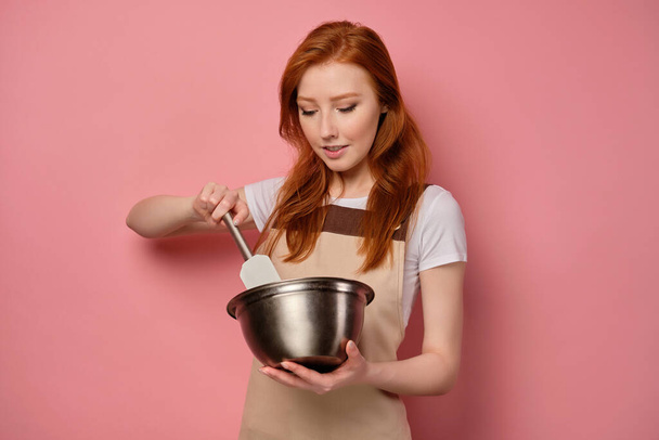 The red-haired girl in an apron stands on a pink background and mixes with a spatula in a bowl - Φωτογραφία, εικόνα