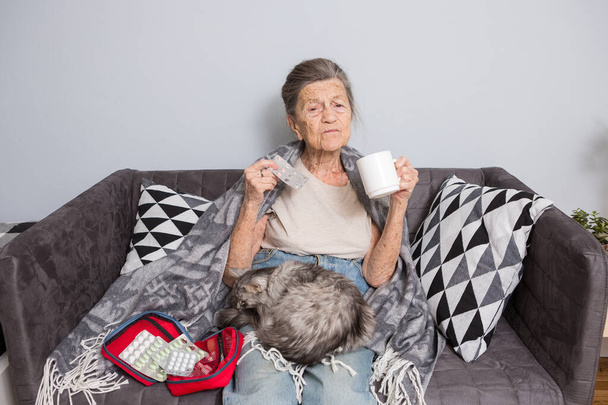 Idős nő, tablettákkal otthon. Egy idősebb nő kezében egy pohár víz és tabletta. A nagyi tablettákat és egy macskát tart a karjaiban a kanapén ülve. Életkor, orvostudomány, egészségügy és az emberek fogalma. - Fotó, kép