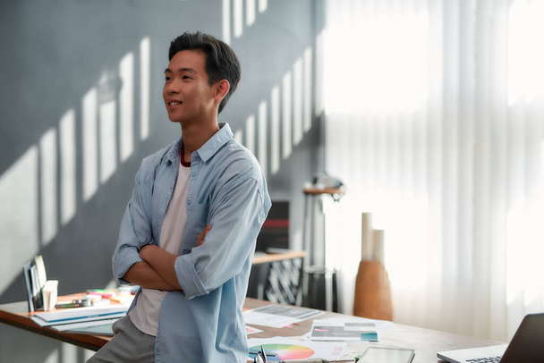 Confident designer. Side view of a young asian man in casual wear with crossed arms looking away and smiling while standing in the modern office - Фото, зображення