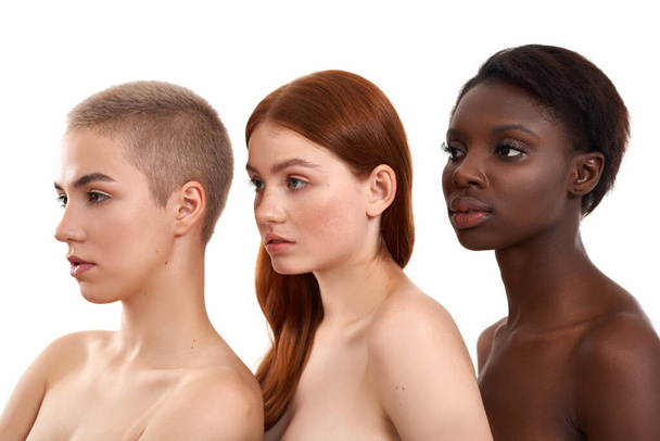 Beauty in Difference. Side view of three beautiful multicultural young women standing close to each other in studio against white background - Fotó, kép