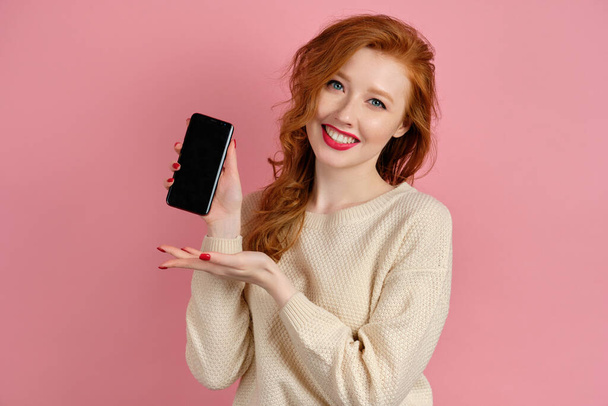 A red-haired girl with red lipstick stands on a pink background pointing at the phone in hand and smiling charmingly - Foto, immagini