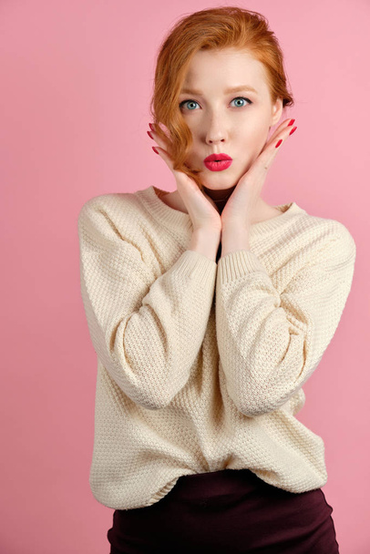 A red-haired girl in a white sweater with red lipstick stands on a pink background holding her hands to her face. - Foto, afbeelding