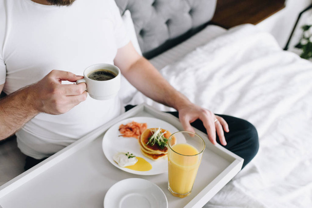 Jonge blanke man eet gezond ontbijt op bed op dienblad - Foto, afbeelding