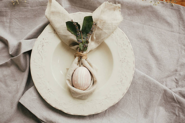 Stylish Easter brunch table setting with egg in easter bunny napkin. Modern natural dyed pink egg on napkin with bunny ears, lavender flowers on vintage plate. Easter table decorations - Фото, изображение