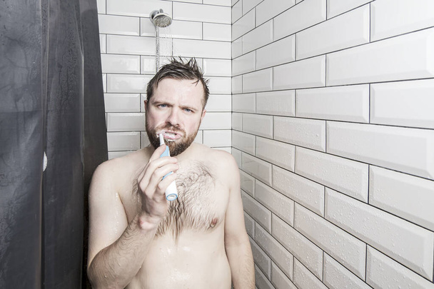 El hombre seguro cepilla los dientes con un cepillo de dientes recargable, de pie en la ducha, bajo el agua corriente y mirando a la cámara
. - Foto, imagen