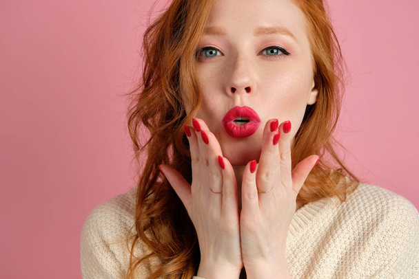 A red-haired curly-haired girl with red lips blows a kiss while looking at the camera, standing on a pink background - Фото, зображення