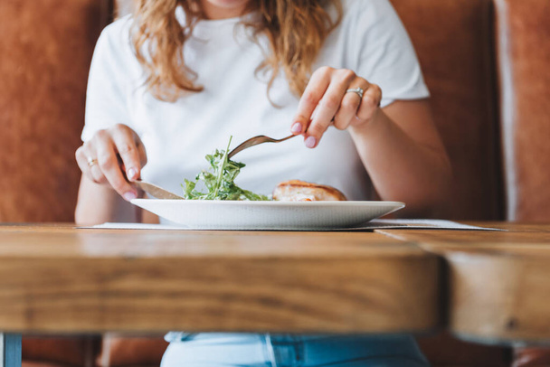 Jovem caucasiana com cabelo encaracolado em camiseta branca comendo salada e frango em um restaurante - Foto, Imagem