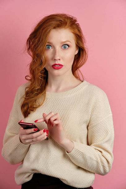 A red-haired girl with red lipstick stands on a pink background with a phone in hands and looks in surprise at the camera - Fotoğraf, Görsel