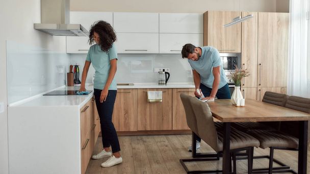 A clean home is a happy home. Two young professional cleaners in uniform working together in the kitchen. Young caucasian man wiping kitchen table, afro american woman cleaning electric stove - Photo, image
