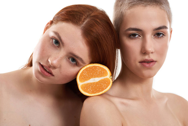 Fruits for a healthy skin. Young red-haired and blonde women holding a piece of orange between their heads while standing against white background - Foto, immagini