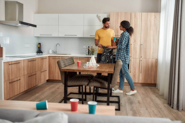 Its almost done. Young multicultural couple in casual clothes holding garbage bag while cleaning their modern kitchen together - Fotografie, Obrázek