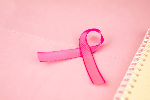 Closeup of pink ribbon lying besides a notebook, isolated in pink cardboard background - Photo, image