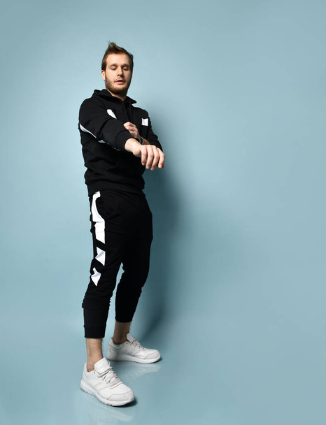 Hipster male in black tracksuit, bracelet and sneakers. He is rolling up his sleeve, posing against blue background. Full length - 写真・画像