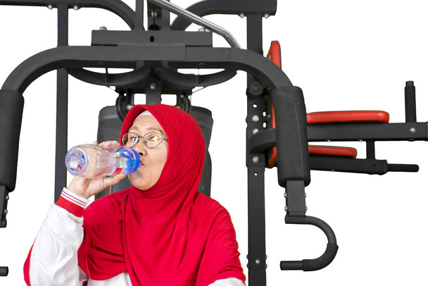 Привлекательная пожилая мусульманка пьет воду, сидя на тренажере после тренировки, изолированная на белом фоне
 - Фото, изображение