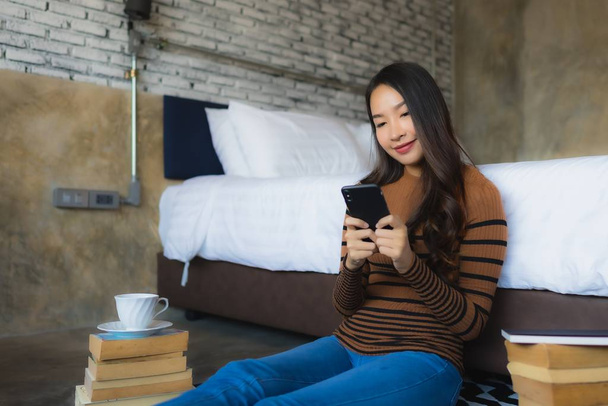 Mladá asijská žena pomocí chytrého mobilního telefonu s čtení knihy a šálek kávy v ložnici interiér - Fotografie, Obrázek