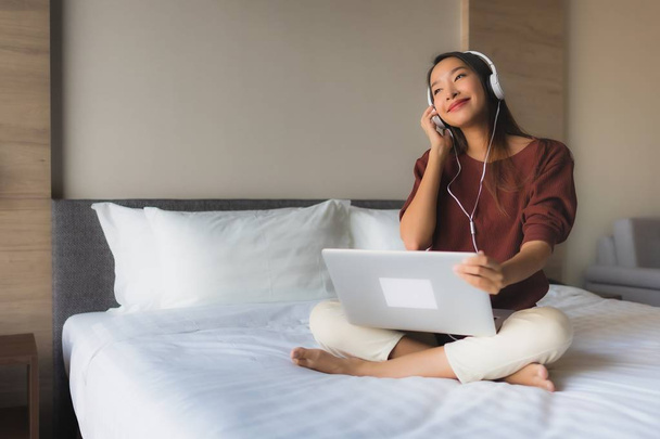 寝室のインテリアでベッドの上で音楽を聴くためにコンピュータとヘッドフォンを使用して美しい若いアジアの女性の肖像 - 写真・画像